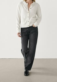 Valkoinen neuletakki, jossa on kultaiset napit ja etutaskut, yhdistetty tummanharmaisiin leveisiin farkkuihin ja mustiin saappaisiin. Sileä, pehmeä rakenne.