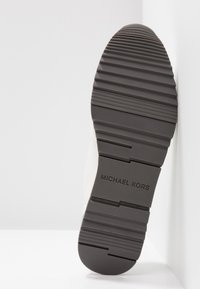 Juodas guminis padas su tekstūruotais briaunomis; ant kulno yra įspausta „MICHAEL KORS”. Šonas yra baltas, demonstruojantis lygų dizainą.