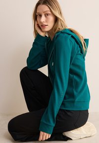 Teal huvtröja i texturerat material, med en framficka och ribbade ärmar. Matchas med svarta byxor och ljusa sneakers.