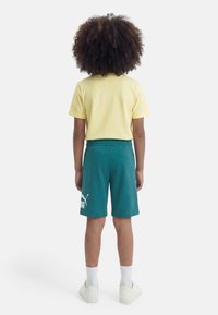 Παιδί με σγουρά μαλλιά στέκεται πλάτη, φορά κίτρινο κοντομάνικο μπλουζάκι, τιρκουάζ σορτς Puma, άσπρες κάλτσες και λευκά αθλητικά παπούτσια.