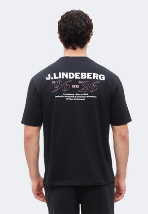 Hombre con cabello rizado que lleva una camiseta negra J.Lindeberg con el texto que celebra 30 años desde 1996 impreso en la parte trasera.