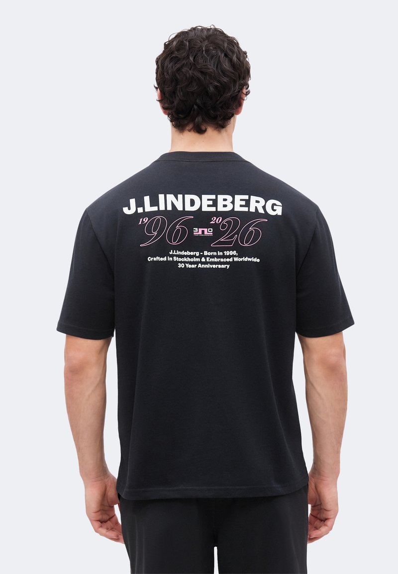 Άνδρας με σγουρά μαλλιά που φοράει μαύρο T-shirt J.Lindeberg με κείμενο που γιορτάζει τα 30 χρόνια από το 1996, τυπωμένο στην πλάτη.