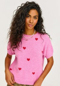 Ružový pletený sveter s krátkymi rukávmi a rozmiestnenými červenými srdiečkami. Mäkka textúra s rebrovaným výstrihom a lemovaním.