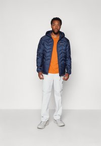 Veste matelassée bleu marine avec capuche, chemise orange en dessous, associée à un pantalon cargo gris clair et des baskets beiges. Design et texture simples.