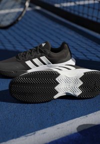 Ασπρόμαυρα παπούτσια τένις Adidas σε γήπεδο μπλε με ρακέτα τένις και δίχτυ στο φόντο.