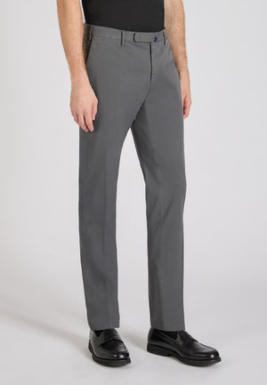 Mann trägt graue gerade geschnittene Hose und schwarze Leder-Loafer, steht vor einem schlichten hellen Hintergrund.