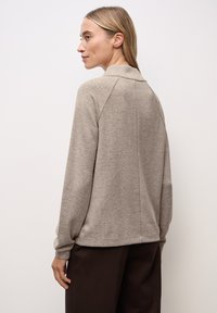 Pull beige avec un col haut et des manches raglan, présentant un tricot texturé et une coupe décontractée, associé à un pantalon marron foncé.