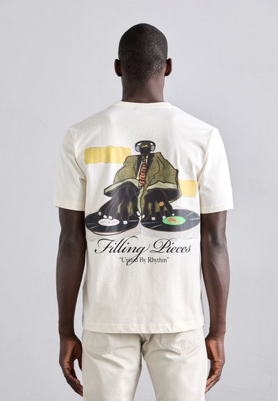 Beige Baumwoll-T-Shirt mit einem Grafikmotiv einer Figur mit Kopfhörern und Schallplatten, auf der Rückseite gedruckt. Der Text beinhaltet "Filling Pieces" und "United By Rhythm."