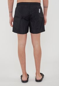 Costumi da bagno neri con finitura testurizzata, vita elastica e due fori con occhielli posteriori. Include un'etichetta con logo sul lato.