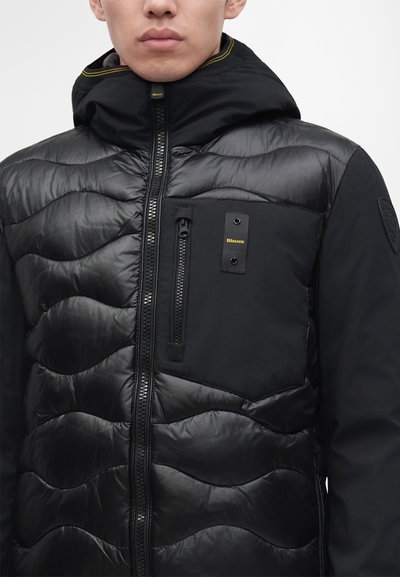 Homme portant une veste matelassée noire avec un col montant, une fermeture éclair devant, une poche poitrine et un petit logo jaune sur la poche.