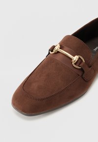 Brune semskede loafers med firkantet tå, forsynet med en gullfarget spenne og svart elastisk detaljering på sidene.