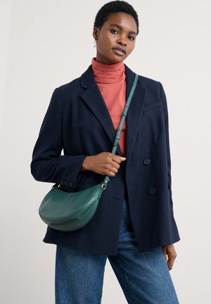 Borsa a spalla in pelle verde con una forma curvata, cuciture dettagliate e tracolla regolabile. Indossata con un blazer navy sopra un dolcevita rosso.