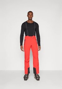 Άνδρας που φοράει μαύρο μακρυμάνικο πουκάμισο, φωτεινά κόκκινο παντελόνι σκι με τιράντες και μαύρα μπότες σκι, στέκεται μπροστά σε ένα απλό λευκό φόντο.