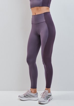 Donna che indossa leggings bicolore viola e scarpe da corsa grigie, in piedi davanti a uno sfondo grigio chiaro uniforme.