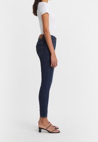 Smala jeans i mörkblå denim med hög midja, klassisk fem-fickorsdesign och en brun läderlogg på baksidan.