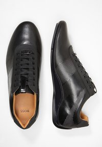 Baskets en cuir noir avec détails perforés, accents brillants et semelle lisse. Intérieur contrastant en cuir tan visible. Design classique à lacets.