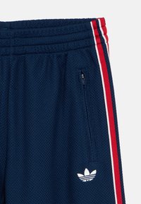 Sportske kratke hlače mornarsko plave boje od teksturiranog materijala, s crvenim i bijelim bočnim prugama, džepom na patentni zatvarač i bijelim logotipom na rubu.