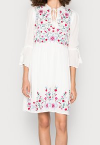 Vestido blanco con bordado floral en rosa, rojo y azul. Presenta un cuello con lazo, mangas acampanadas y una falda fluida con un dobladillo decorativo.