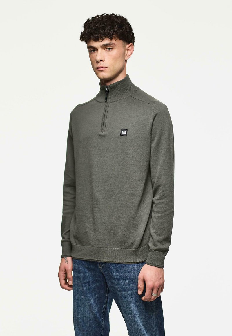 Mann in einem olivgrünen Pullover mit Viertelreißverschluss und blauen Jeans, der vor einem schlichten weißen Hintergrund steht.
