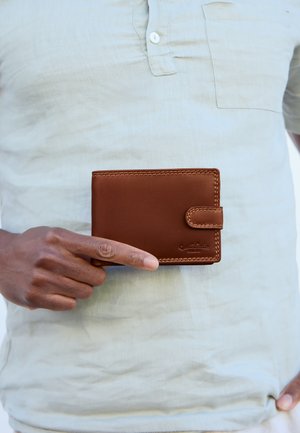 Personne portant une chemise beige tenant un portefeuille en cuir marron avec une fermeture pression et des bords cousus au niveau du torse.