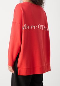 Röd oversized sweatshirt gjord av mjukt tyg, med rund hals, långa ärmar och ett vitt logotryck på baksidan. Sidoflikar i kanten.