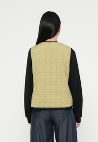 Prošívaná vesta v světle zelené barvě s trojúhelníkovými vzory, spojená s černým tričkem s dlouhým rukávem, pohled zezadu. Volný střih, neformální design.