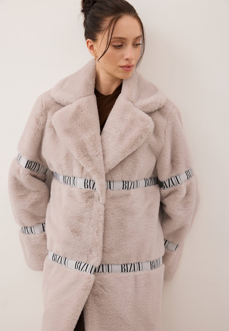 Jasnoróżowy płaszcz z futerka sztucznego z głębokim kołnierzem, oversize'owymi rękawami i białymi pasami z napisem „BIZUU” wokół talii i rękawów.