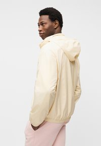 Νέος άνδρας που φοράει ανοιχτόκίτρινο μπουφάν με κουκούλα και ανοιχτό ροζ παντελόνι, στέκεται πλαγίως με τα χέρια στις τσέπες μπροστά σε απλό φόντο.