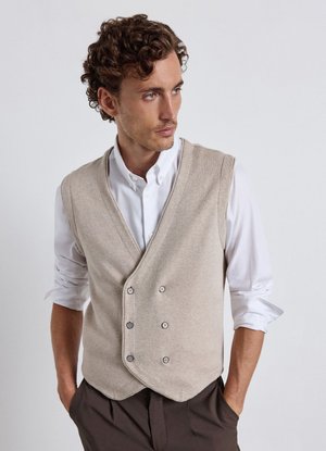 Man with curly hair wearing a beige buttoned vest over a white shirt with rolled-up sleeves and dark pants, hands in pockets, looking sideways.