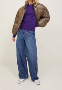 Doudoune marron, pull violet, jean bleu ample, baskets blanches. La doudoune présente une fermeture éclair à l'avant et des poignets côtelés.