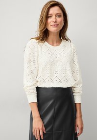 Pull en tricot blanc avec un motif floral en 3D et un col rond. Manches longues et coupe courte. Associé à une jupe en cuir noir ajustée.