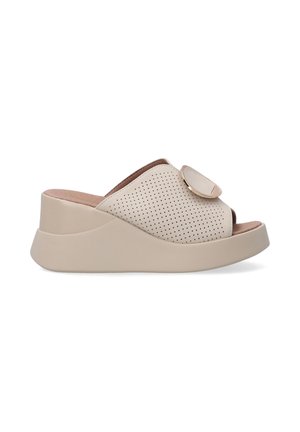 Beige perforated slip-on shoes with a smooth sole, featuring a rounded toe and a decorative circular accent on the upper.