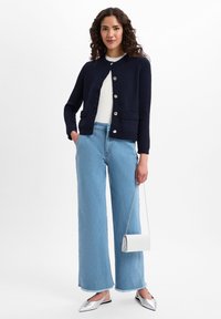 Cardigan en maille marine avec fermeture à boutons, jean bleu clair à jambes larges, ballerines argentées à bout pointu et un petit sac à main blanc avec une bandoulière en chaîne.