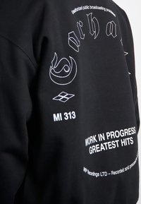 Felpa nera con testo e grafiche ricamate bianche, inclusi "WORK IN PROGRESS", "MI 313" e motivi decorativi. Texture morbida.