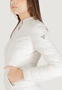 Giacca imbottita bianca con colletto alto, chiusura con zip e tasche inclinate. Presenta una toppa con logo triangolare sulla manica. Tessuto morbido.