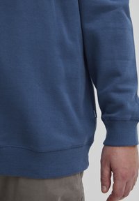 Sudadera azul con puños y dobladillo acanalados. Textura suave, diseño casual, con una pequeña etiqueta de la marca cerca del dobladillo en el lado izquierdo.
