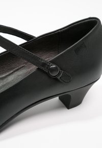 Chaussure en cuir noir avec un petit talon, présentant un design à brides croisées et un détail de fermeture par bouton. Texture lisse, silhouette élégante.