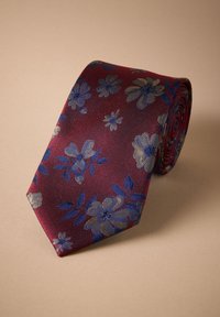 Cravate rouge foncé roulée avec des motifs floraux bleus et dorés sur une surface beige clair.