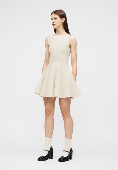 Νέα γυναίκα με ίσια καστανά μαλλιά, φορούσα αμάνικο εκρού φόρεμα με φούστα σε φαρδιά γραμμή, λευκές κάλτσες και μαύρες γόβες Mary Jane, στέκεται σε λευκό φόντο.