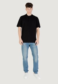 Camisa polo negra de manga corta con una tapeta de dos botones, combinada con jeans de color azul claro y zapatillas deportivas blancas. El modelo se encuentra de pie frente a un fondo neutro.