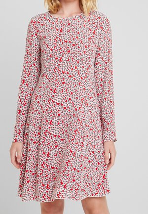 Mujer con un vestido de manga larga y largo hasta la rodilla, con un pequeño estampado floral en rojo, negro y blanco sobre un fondo claro.
