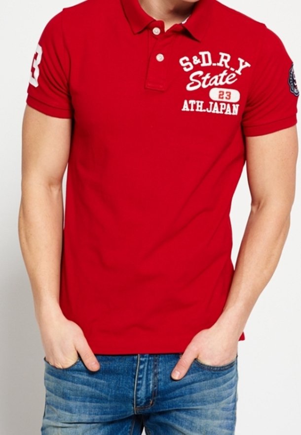 Homme portant un polo rouge avec un texte blanc "S&DRY State ATH Japan 23" et un jean bleu, les mains dans les poches.