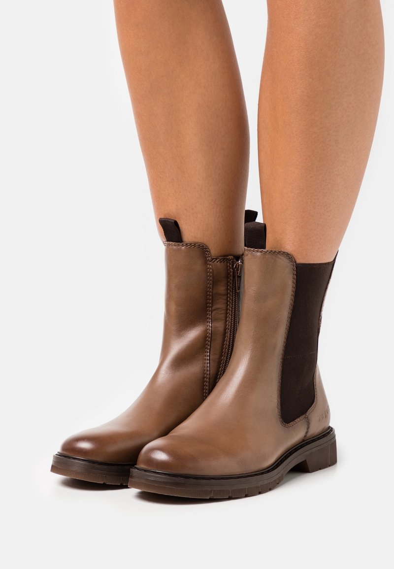 Bottines en cuir marron avec fermeture éclair sur le côté et panneaux élastiques, portées sur des jambes nues sur un fond blanc.