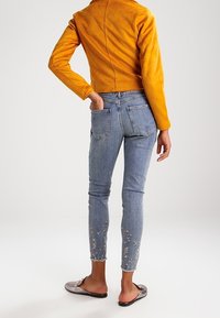 Gul mocka kort jacka, ljusblå denimjeans med blommig broderi vid vristerna, och silverfärgade platta slip-on skor.