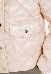 Gepolsterte, hellbeige Jacke mit einem Rautenmuster, Druckknöpfen und einer großen Vorderasche. Glatte, glänzende Stoffoberfläche.