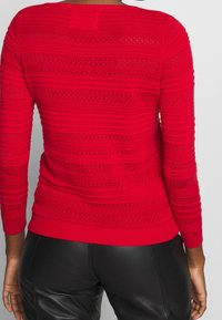 Maglione rosso in maglia costine con strisce orizzontali e dettagli traforati, maniche lunghe. Design aderente con scollo rotondo.