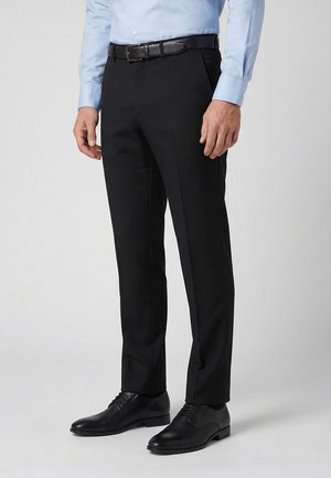 Mand iført sorte habitbukser, sort bælte, sorte pæne sko og en lyseblå skjorte med lange ærmer, stående foran en ensfarvet baggrund.