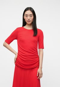 Κόκκινη μπλούζα κοντομάνικη από λεία ύφανση, με ραβδώσεις στα πλάγια και στρογγυλή λαιμόκοψη, συνοδευόμενη από μία ταιριαστή φούστα.