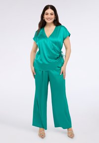 Zielona satynowa bluzka zapinana z krótkim rękawem, z detalem w postaci kokardy z boku, zestawiona z szerokimi spodniami w tym samym kolorze. Widoczne srebrne sandały.