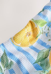 Tissu à texture gaufrée avec des rayures bleues et blanches, des tranches de citron jaune et des feuilles vertes, avec un petit emblème métallique rond.